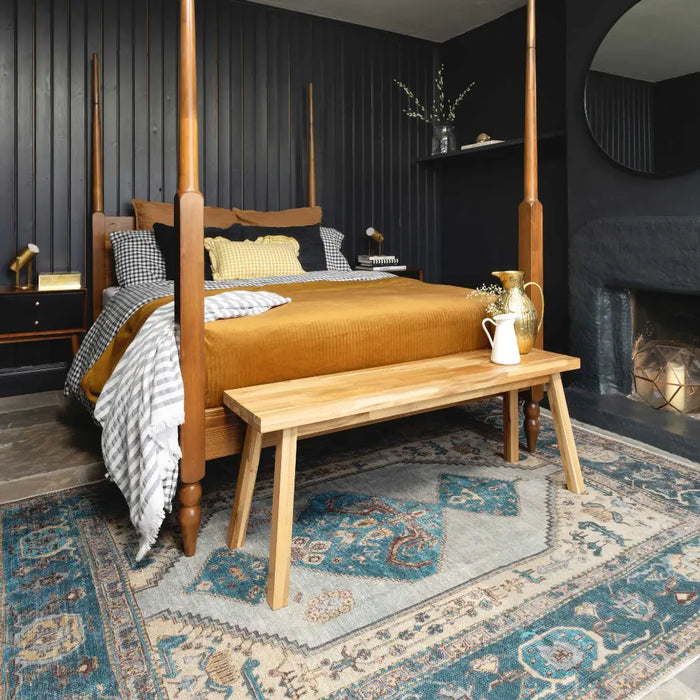 Large wooden four posted bed sits stop a blue Persian rug. The bed has a mustard yellow blanket on top and lots of cushions. The walls are black wooden panelling and a fire is featured at the right hand side of the image.