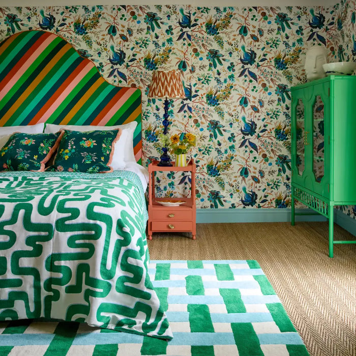 A blue and green checked rug features in the middle of a colourful bedroom with a large bed with patterned duvet and striped headboard. In the background, the walls are covered with colourful, floral wallpaper.