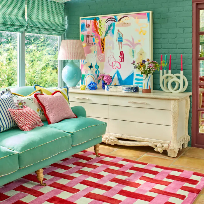 A bright pink checked rug features in the centre of a living room with striking turquoise walls and large windows. Atop the rug sits a matching green sofa with colourful cushions. There is a white sideboard with many decorations.