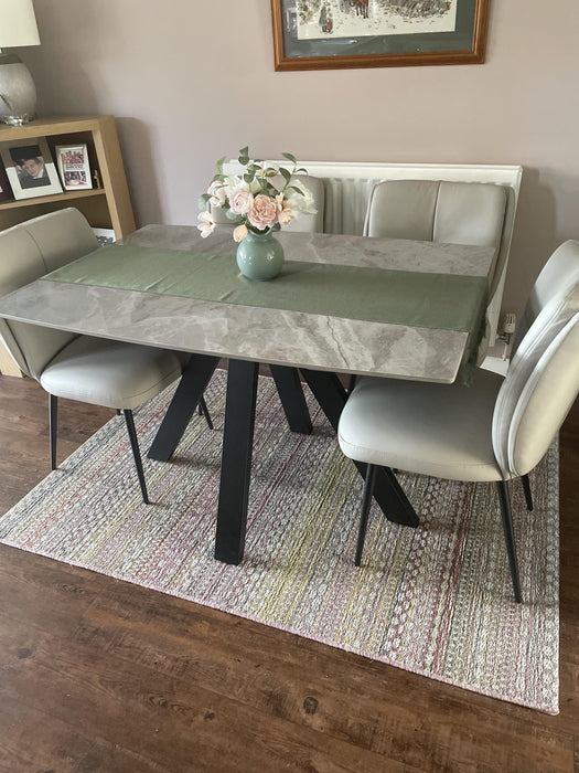 Photo By Carol L | A modern dining table set in a dining room, featuring a gray marble-patterned top, black angled legs, and a green table runner. It is surrounded by four light gray upholstered chairs on a patterned rug over dark wood flooring.