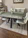 Photo By Carol L | A modern dining table set in a dining room, featuring a gray marble-patterned top, black angled legs, and a green table runner. It is surrounded by four light gray upholstered chairs on a patterned rug over dark wood flooring.