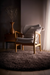A modern wooden armchair with light gray upholstered seat and back, styled with a striped throw pillow, set on a round dark brown shag rug in a dimly lit room with a side table and vase.