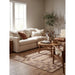 A beige upholstered sofa in a styled living room with neutral-toned walls, a round wooden coffee table, a patterned area rug, and a potted tree.